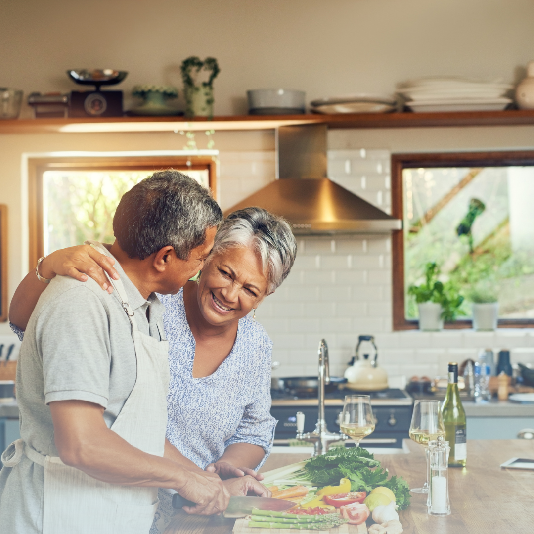 An elderly woman smiling with her hand over the shoulder of an elderly man. The two are standing in a kitchen and are next to a counter with fresh vegetables.