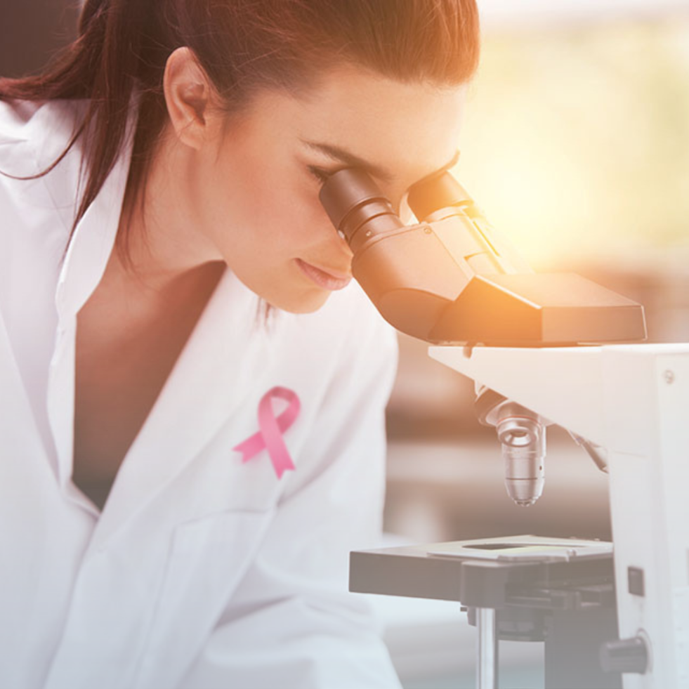 Woman in white lab coat with a pink ribbon pin looks through a microscope.