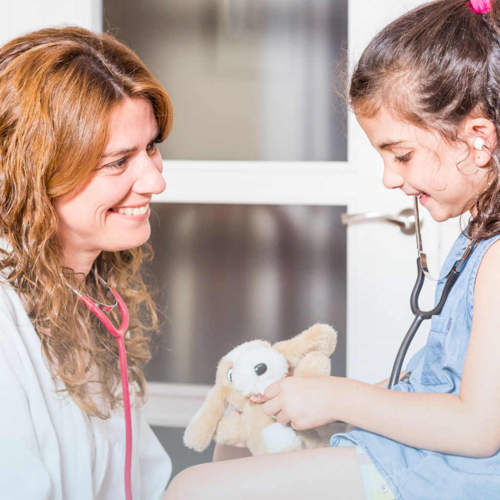 Woman with red hair and a red stethoscope smiling at a young girl with brown hair who is hold a teddy bear while smiling.
