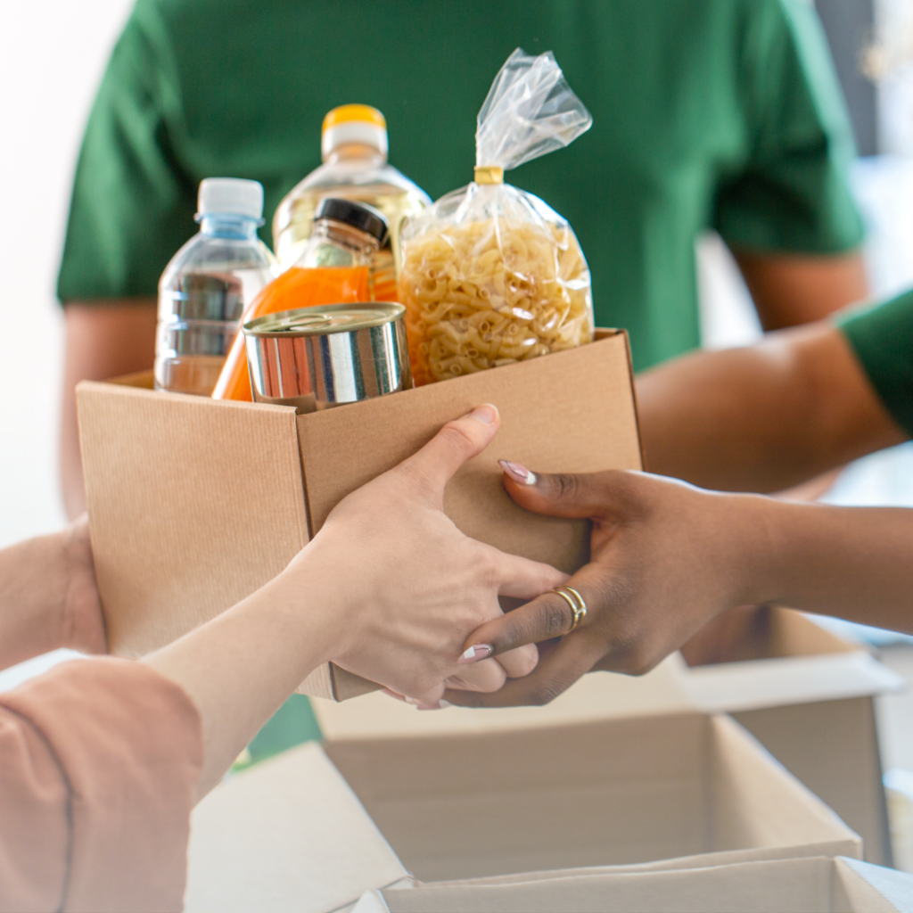A cardboard box of canned food, water and snacks being exchanged between two people whose arms are only visible.