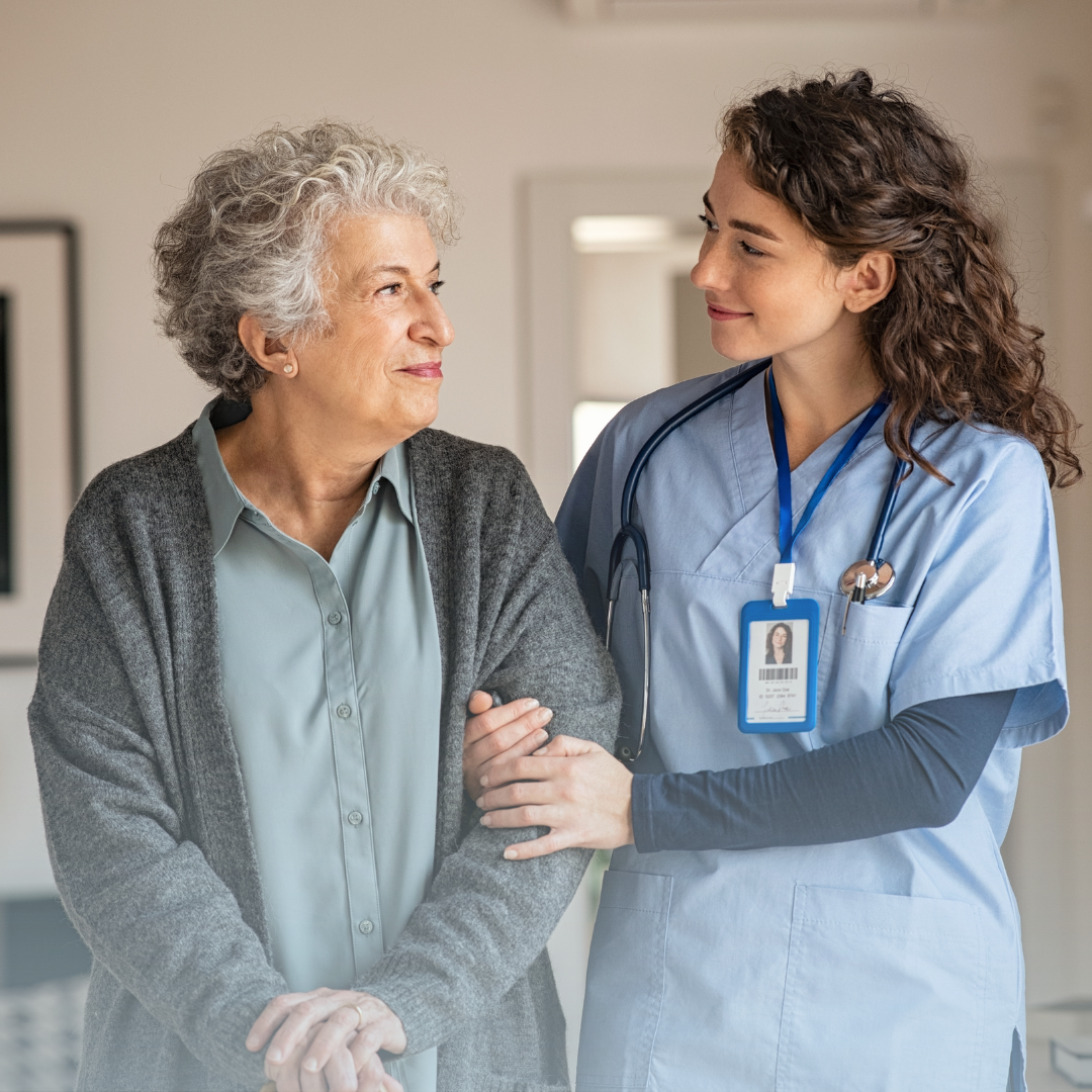 Female nurse on the right holding on to the arm of an elderly resident on the left. Both are smiling looking at eachother.