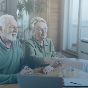 An elderly, white couple, both with white hair and in green tops, are sitting at a table, smiling. The couple is a man (on the left) and a woman (on the right). The woman is shaking the hand of a younger white woman, who is across from them. That woman is out of frame.