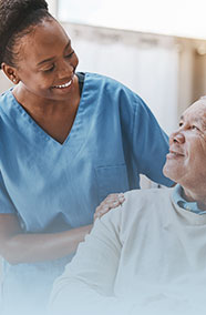 An elderly Black man in a grey sweater sits in a wheelchair and is looking up at a Black female nurse in blue scrubs who is next to him on his right. They are both smiling and her hands are on this shoulders in a comforting manner.