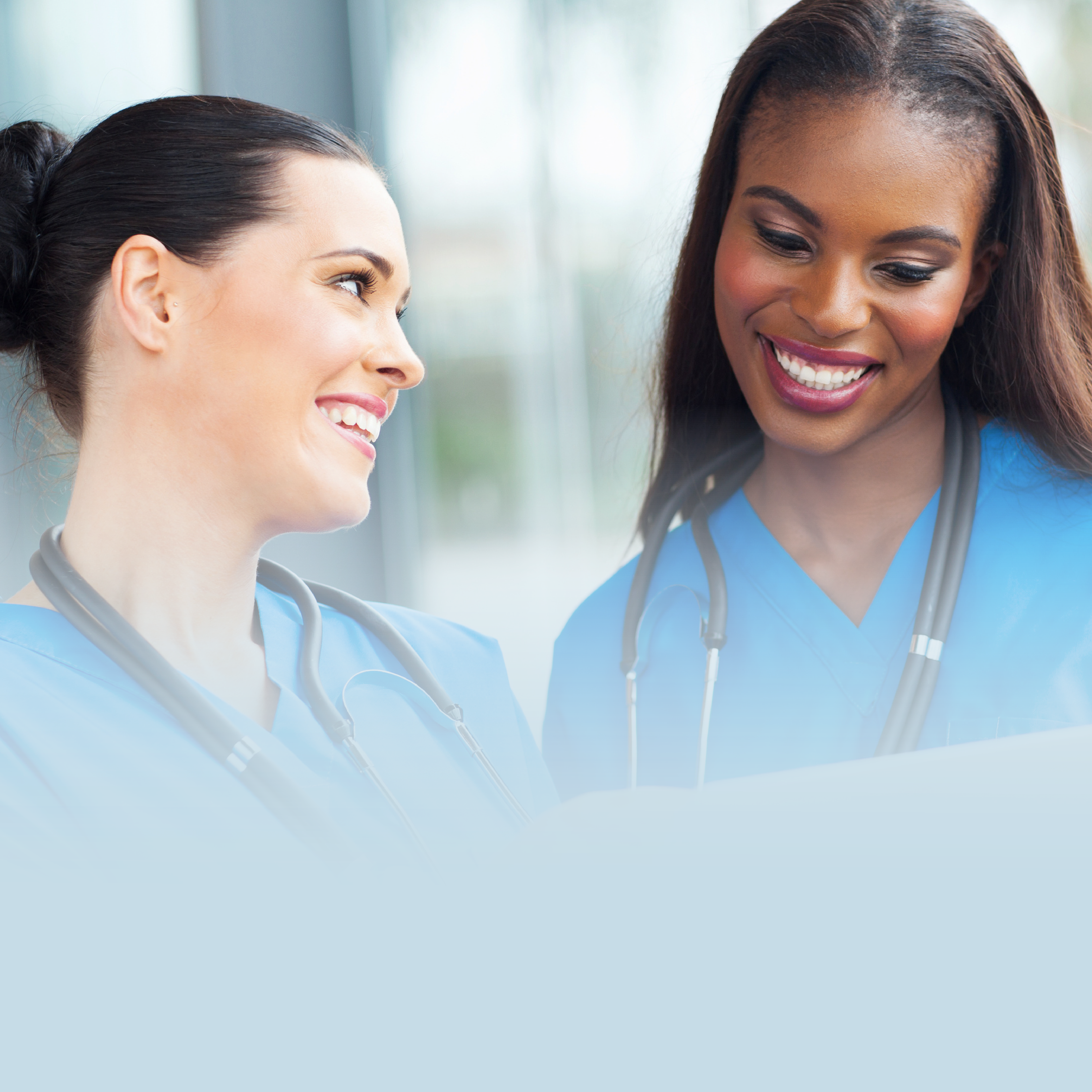 The image shows two young female nurses smiling. The one on the left is white, with black hair that is tied back in a bun. She is smiling and looking at her colleague to her right, who is Black with dark brown hair that is worn down. She is smiling at the ground. They are both wearing light blue scrubs with stethoscopes around their necks.