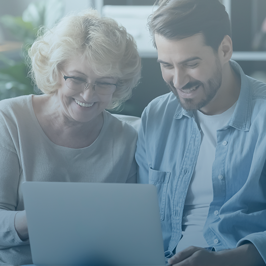 A mother and son sit together, smiling in front of a computer. The woman on the left is elderly, with white hair and wearing a cream sweater. The man on the right is middle-aged and has brown hair. He is wearing a blue buttoned-down shirt with a white T-shirt underneath.