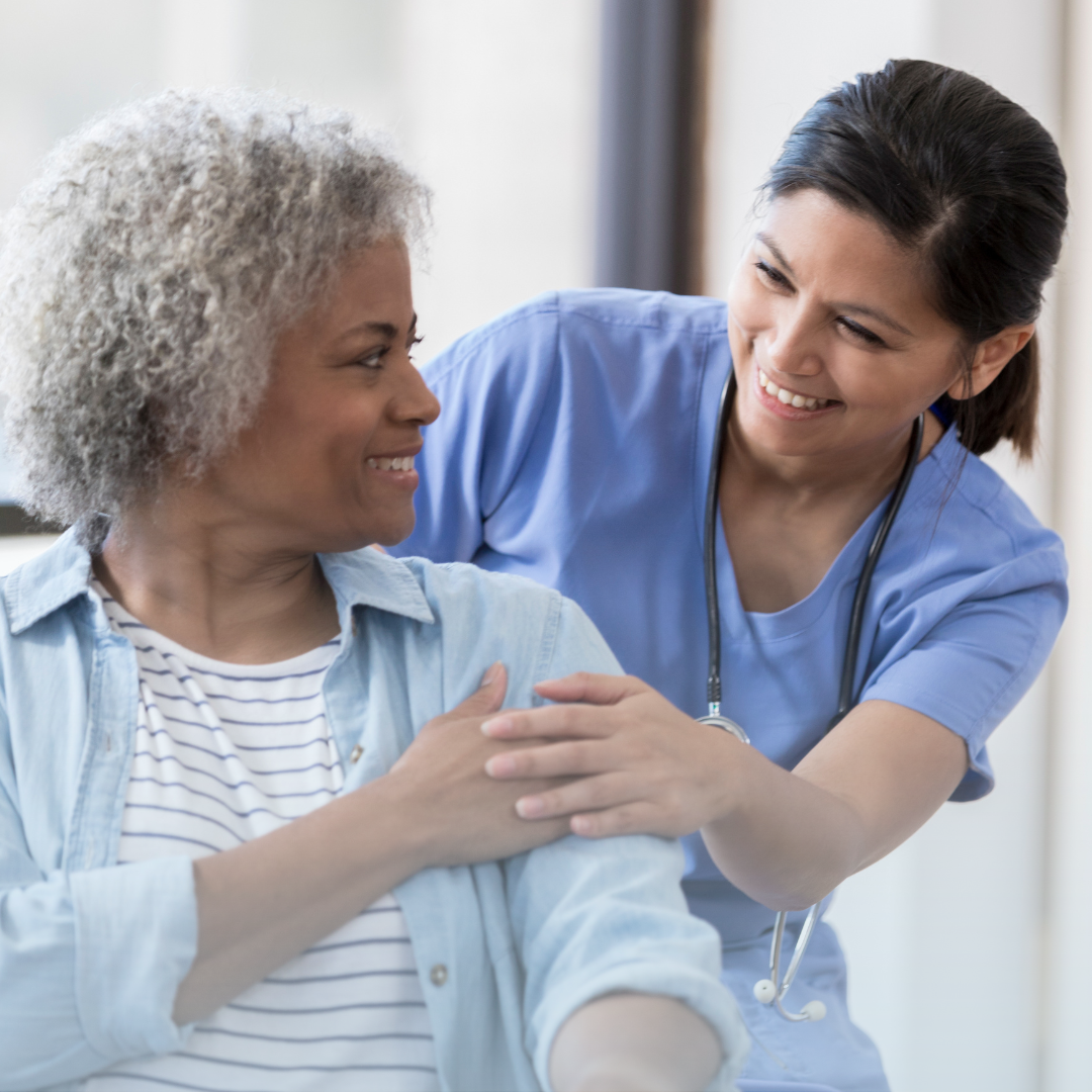 Female nurse in a blue uniform smiling as she touches the arm of a patient.